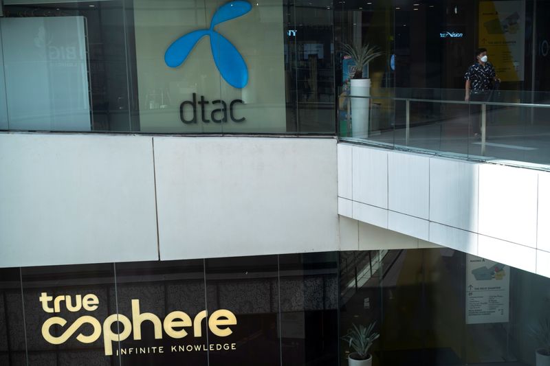FILE PHOTO: Man walks next to logo of the DTAC Public Company Limited and logo of True Corp. in Bangkok