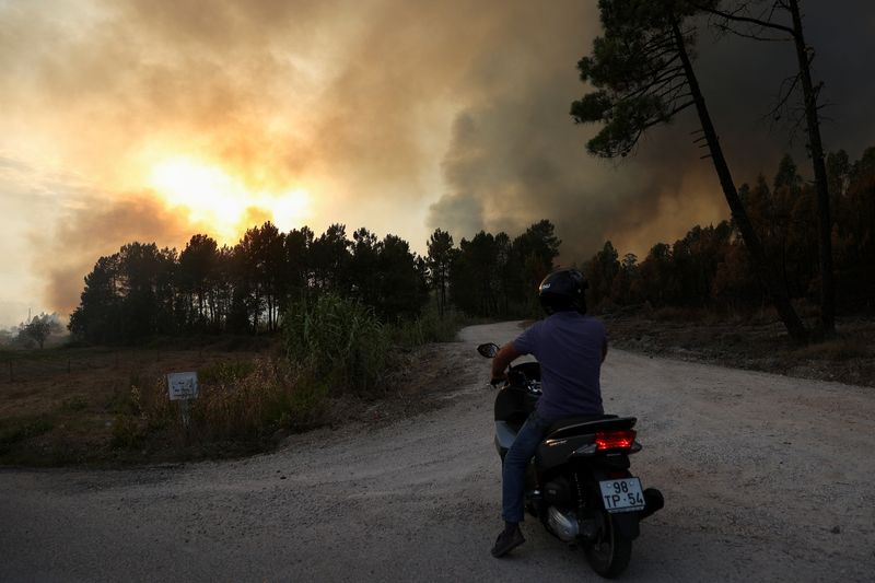 A person sits on a motorcycle as smoke rises from a wildfire in Ourem