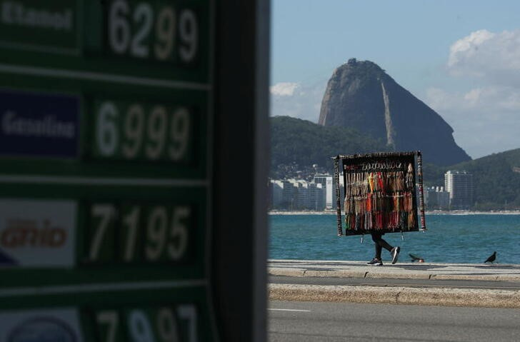 Imagen de archivo de un vendedor callejero andando por la playa de Copacabana junto a una gasolinera con los precios expuestos al público en Río de Janeiro, Brasil.