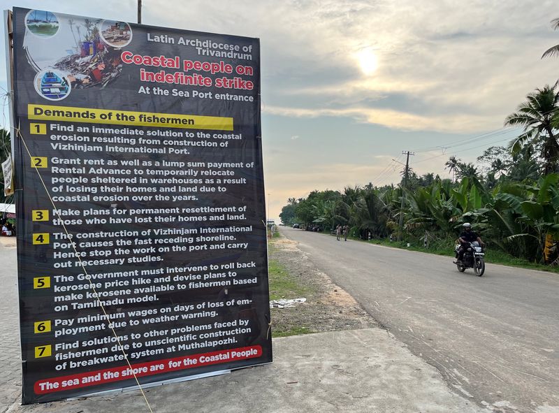 A motorcyclist moves past a placard featuring the demands of protesting fishermen near the entrance of the proposed Vizhinjam Port